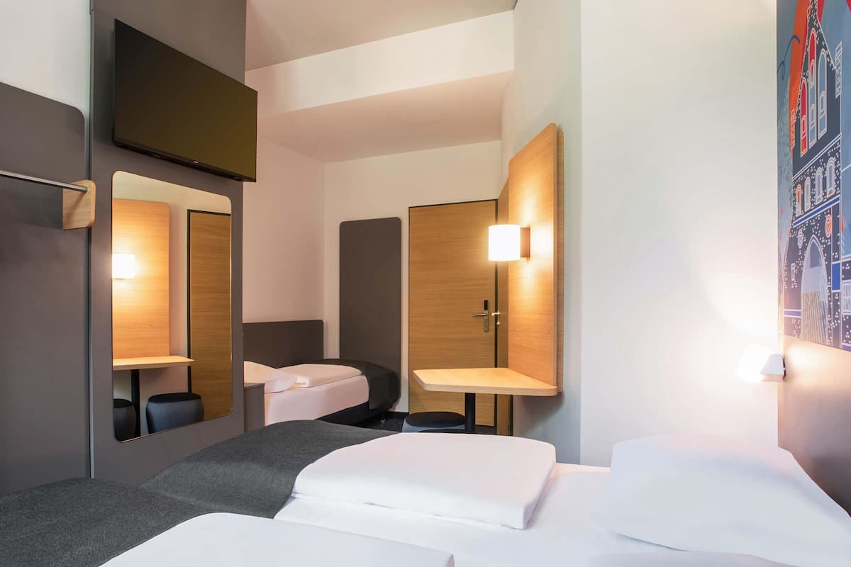 A hotel room with two beds, a TV, a mirror, a bedside table, and a wall-mounted lamp.