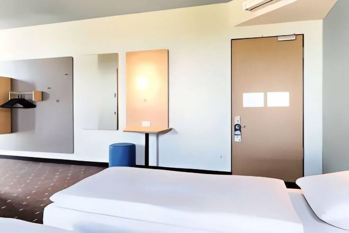 A hotel room with a bed, a desk, a chair, a lamp, a door, and a wall-mounted mirror.