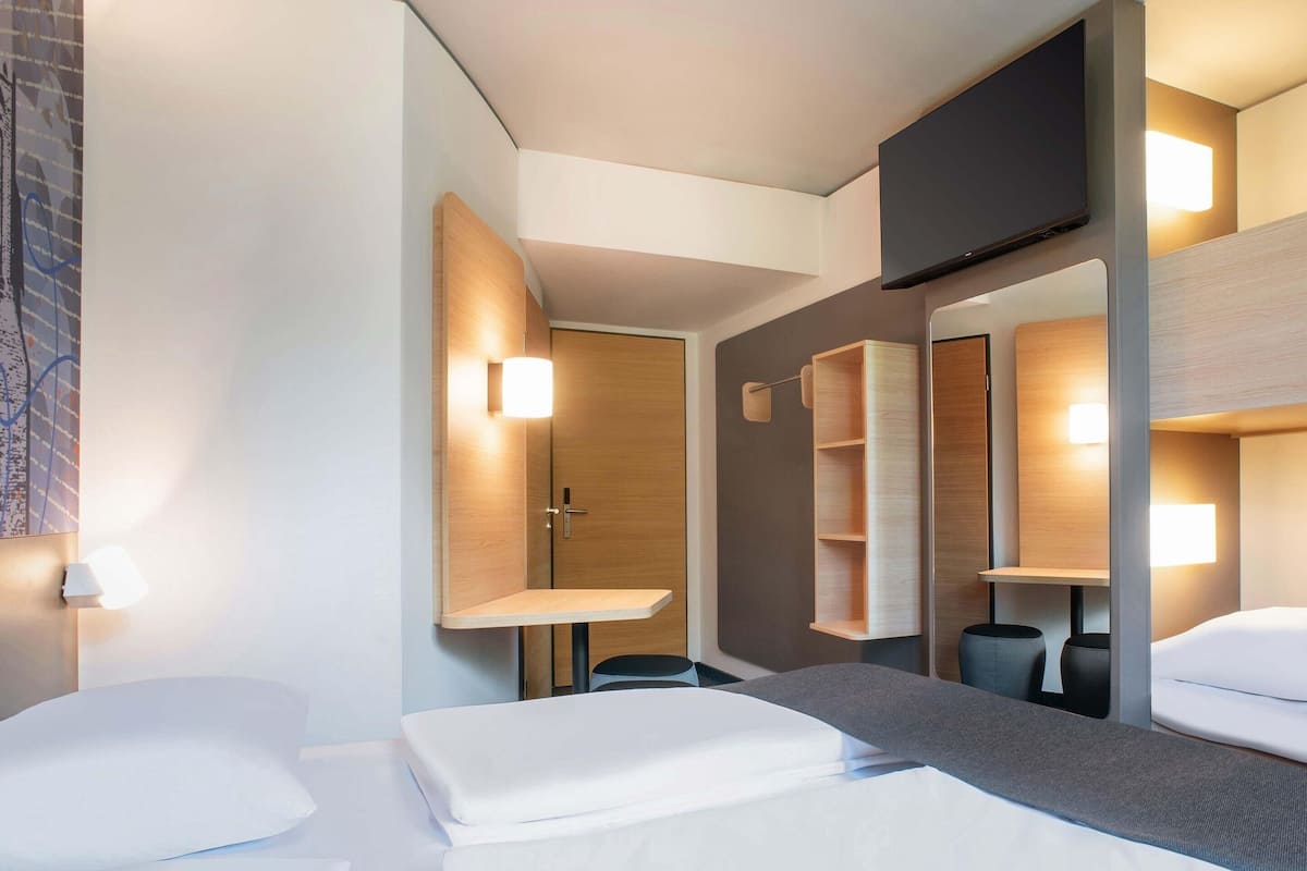 A hotel room with a bed, a TV, a mirror, and a wall-mounted shelf.