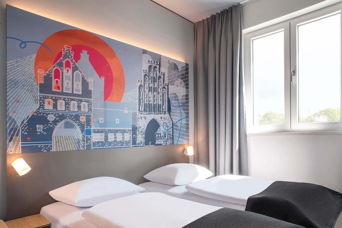 A hotel room with two beds, a large wall mural, a window with curtains, and a nightstand with a lamp.