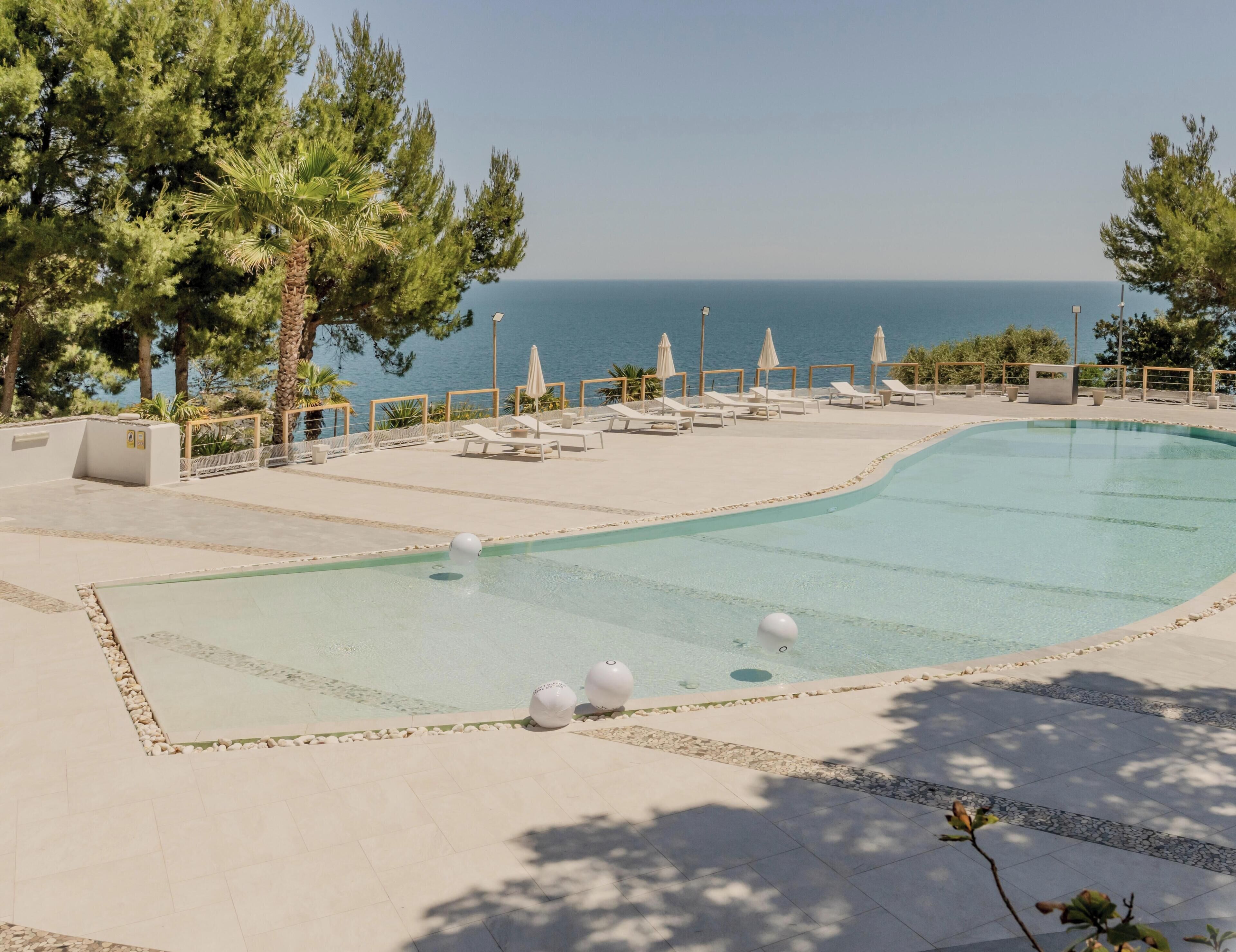 A swimming pool with lounge chairs and umbrellas, surrounded by trees and a view of the sea.