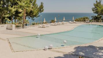 A swimming pool with lounge chairs and umbrellas, surrounded by trees and a view of the sea.