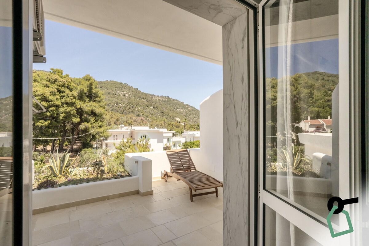 A balcony with a wooden lounge chair, a view of a hillside with buildings, and a clear sky.