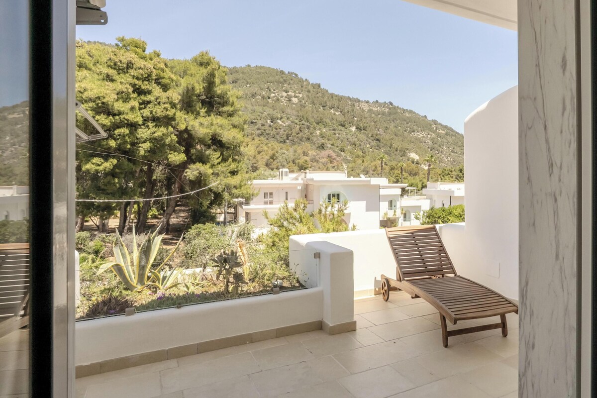 A balcony with a wooden lounge chair, a view of a hillside with buildings, and a clear sky.