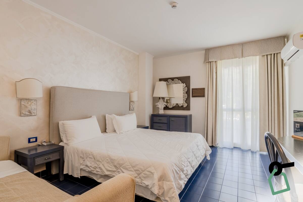 Quarto de hotel com uma cama grande, mesas de cabeceira, uma escrivaninha e uma janela com cortinas.