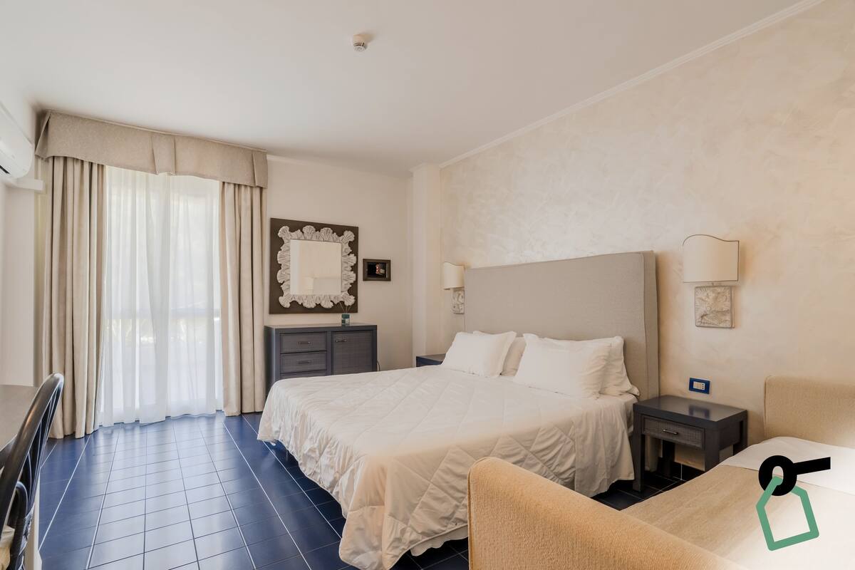Quarto de hotel com uma cama grande, uma mesinha de cabeceira com abajur, uma cadeira e uma escrivaninha.