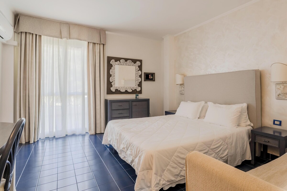 A hotel room with a large bed, a bedside table with a lamp, a chair, and a desk.