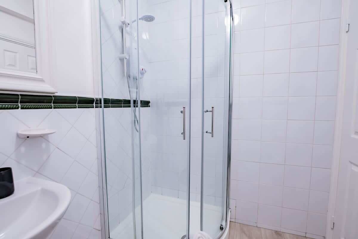 A glass-enclosed shower with a white tiled wall and a white sink to the left.