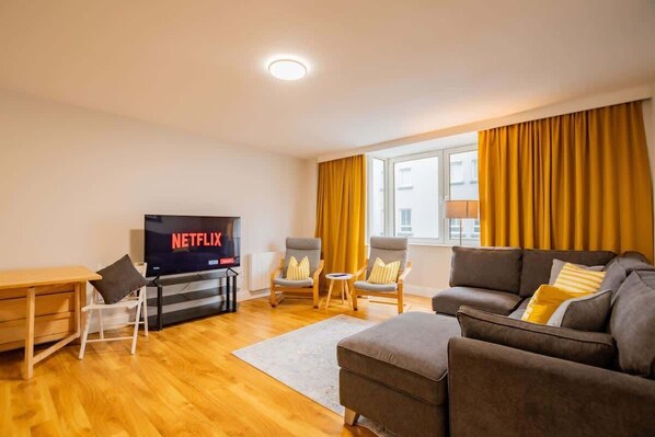 Een moderne woonkamer met een flatscreen-tv waarop 'NETFLIX' wordt weergegeven, een bank, fauteuils, een houten salontafel en een bijzettafeltje.