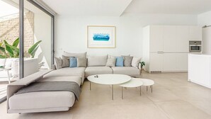 A modern living room with a sectional sofa, two small tables, and a kitchen area with white cabinets and appliances. - 4-bedroom modern villa with private pool and spectacular views! (La Caleta)