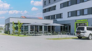 Un hôtel moderne avec une façade en verre, un espace extérieur aménagé pour s’asseoir et un fourgon blanc garé devant.