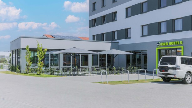 Un hotel moderno con fachada de vidrio, zona de estar al aire libre y una furgoneta blanca estacionada frente a él.
