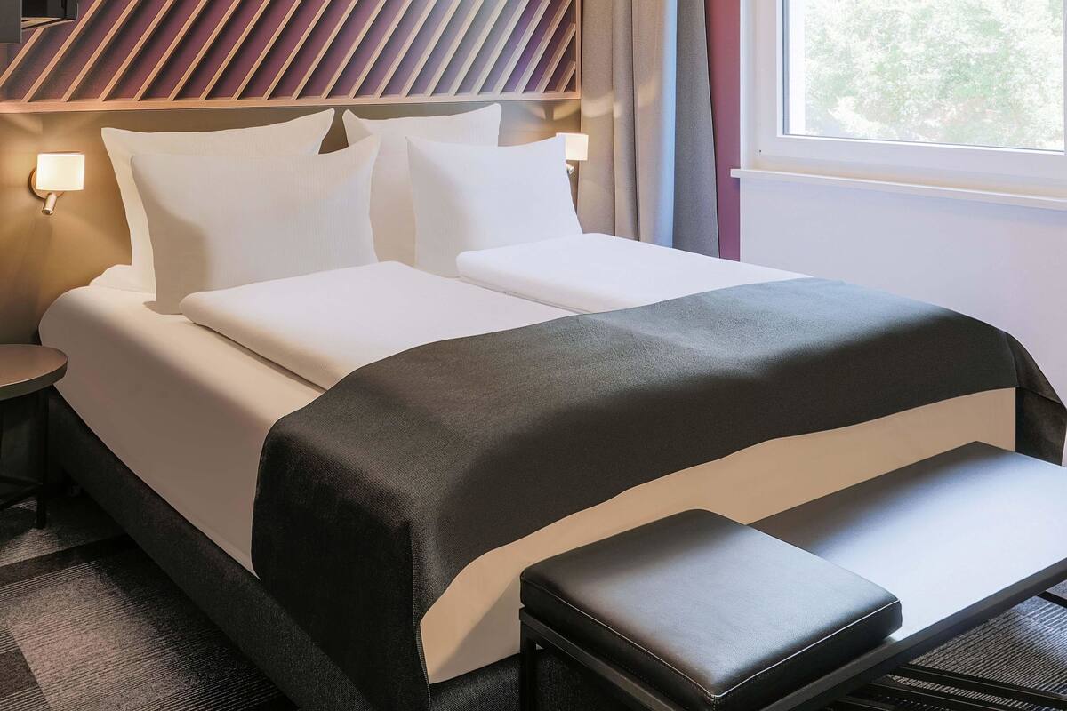 Une chambre d’hôtel avec un lit, des tables de chevet, un banc et une fenêtre avec des rideaux.