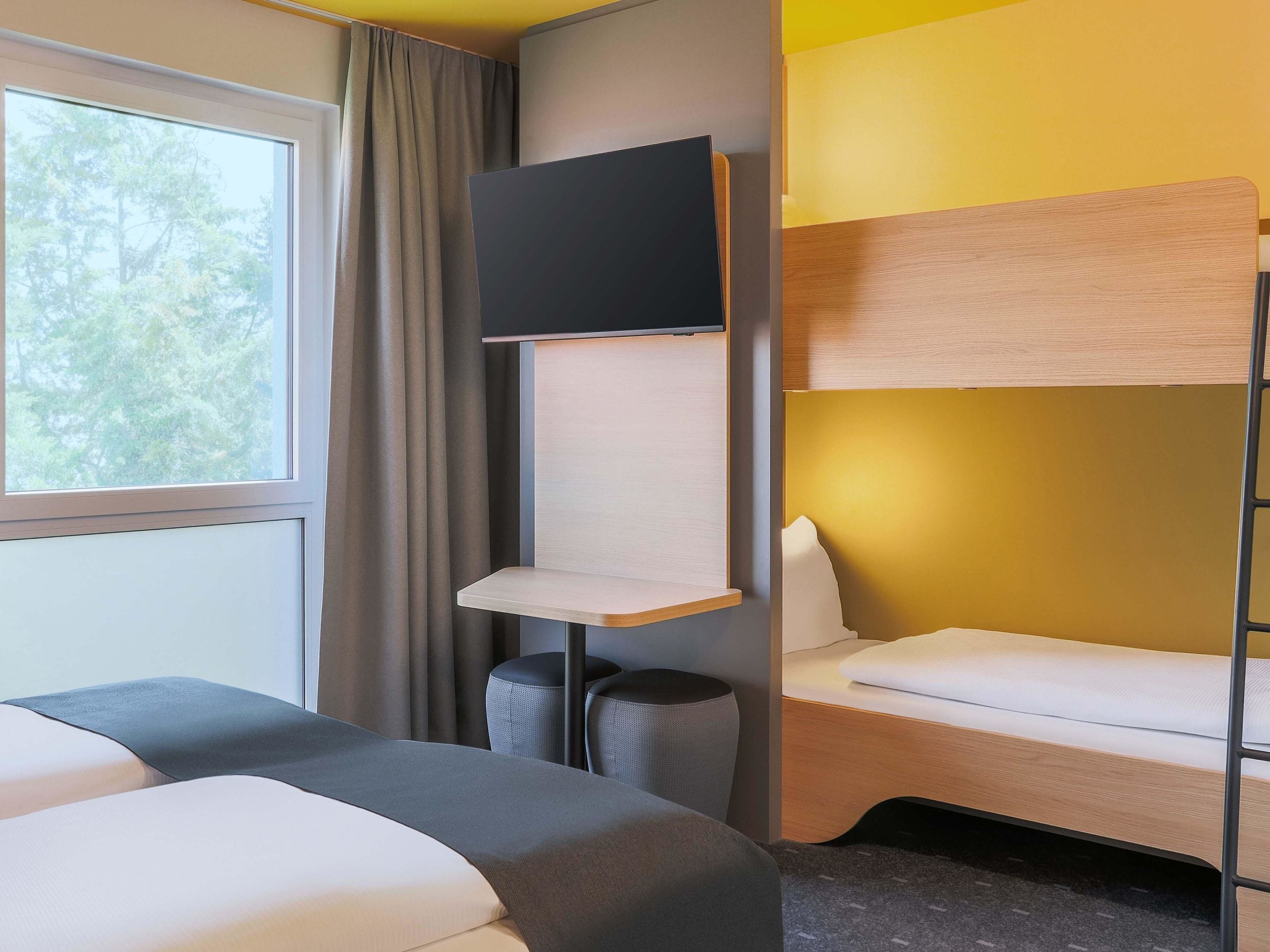 Una habitación de hotel moderna con una cama litera, un televisor de pantalla plana montado en la pared, una mesita de noche y una ventana con cortinas.