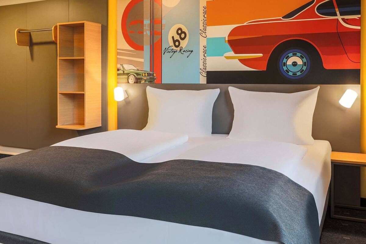 Une chambre d’hôtel avec un lit, une table de chevet, une étagère et un mur orné d’une affiche de voiture de course.