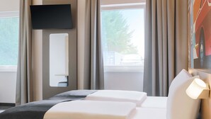 Une chambre d’hôtel moderne, équipée d’un lit, d’une table de chevet, d’un téléviseur fixé au mur et d’une fenêtre avec des rideaux.