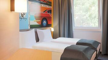 A hotel room with a bed, a small table, a window with curtains, and a wall with a racing car poster.