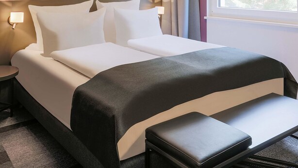 Une chambre d’hôtel avec un lit, des tables de chevet, un banc et une fenêtre avec des rideaux.