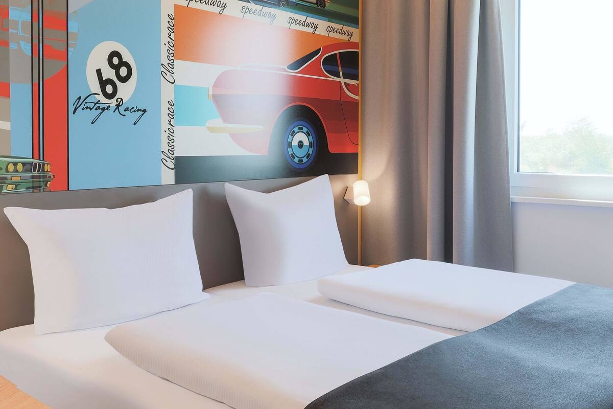Une chambre d’hôtel avec un lit, des oreillers, une table de chevet, une lampe et une fenêtre avec des rideaux.