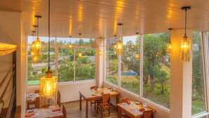 A dining area with tables and chairs, large windows with a view of greenery, and hanging pendant lights. - Mount Hunt Resort (Beragala)