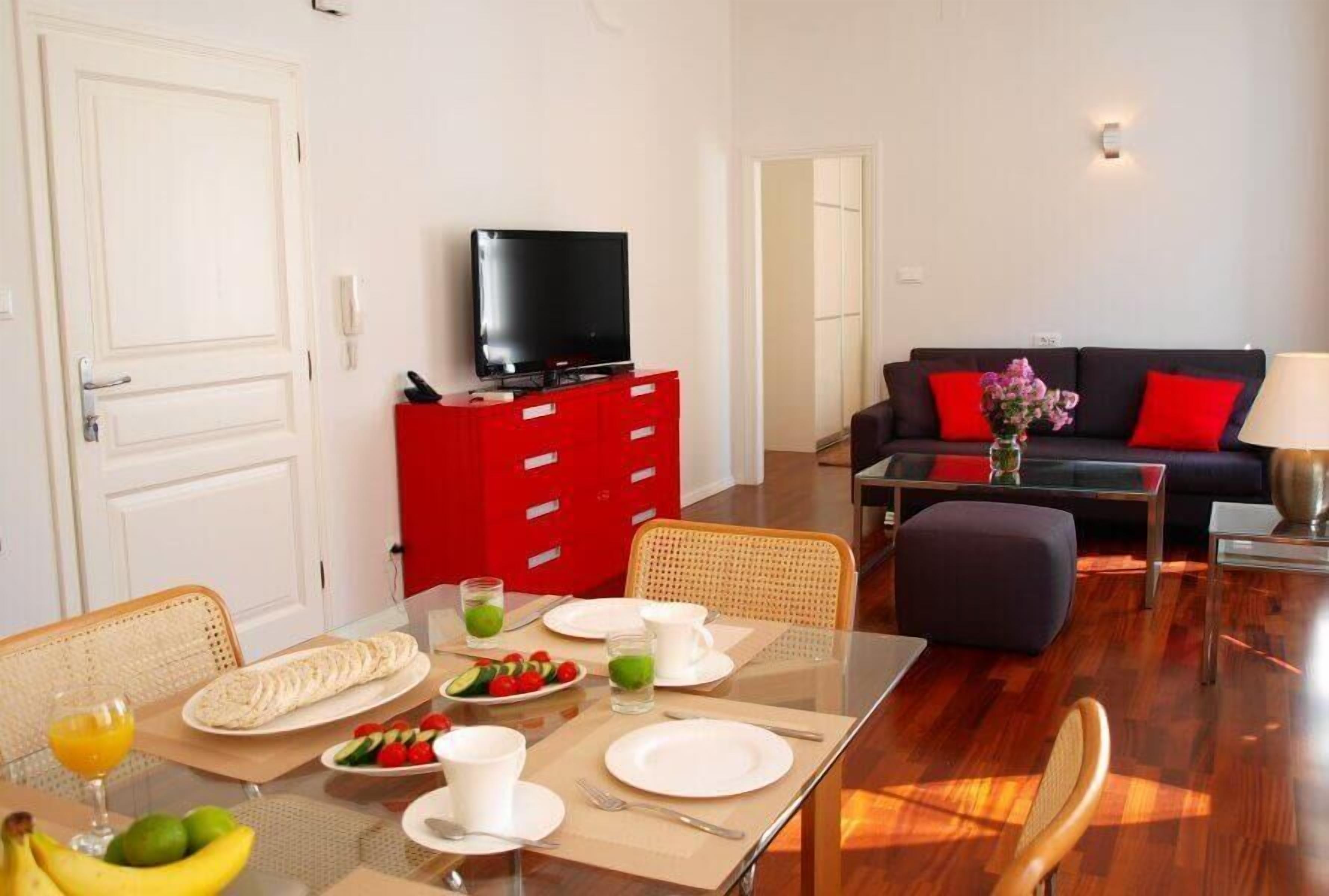 A dining area with a table set for two, a red cabinet with a television, and a living area with a sofa and red cushions.