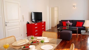 A dining area with a table set for two, a red cabinet with a television, and a living area with a sofa and red cushions. - La Dolce Vita-Old Town (Dubrovnik)