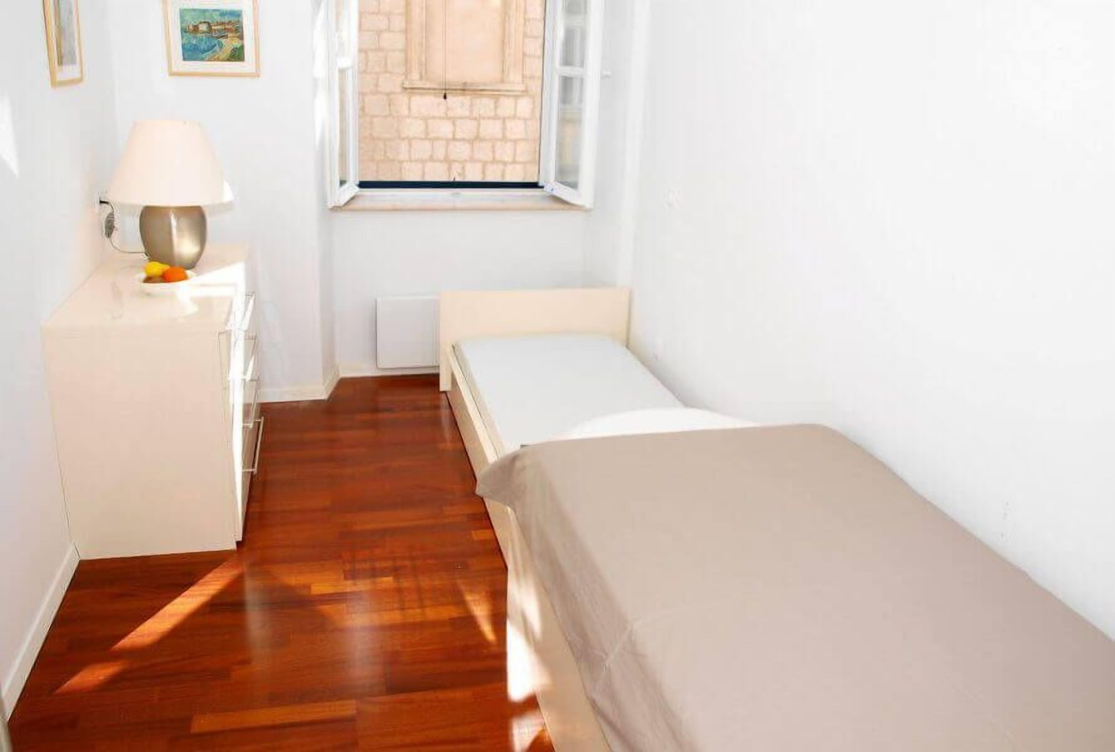 A small, bright room with a wooden floor, a single bed, a nightstand with a lamp, and a window with a view of a brick wall.