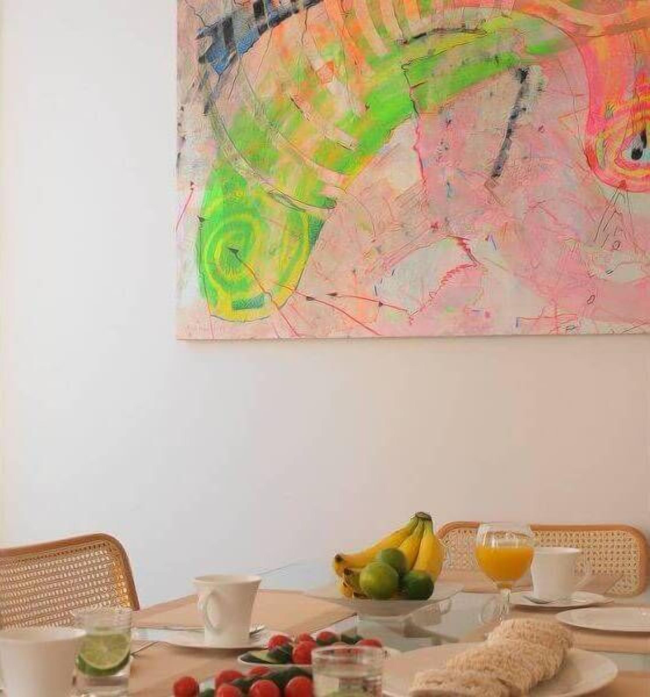 A dining area with a table set for breakfast, including fruits, a glass of orange juice, and a cup. A colorful abstract painting is on the wall above the table.