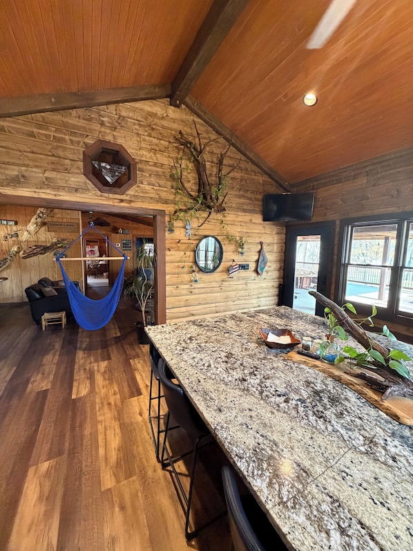A rustic interior with a wooden ceiling, a hammock, a bar area with stools, and a large mirror.
