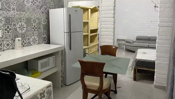 A compact kitchen with a refrigerator, microwave, and a patterned backsplash. There is a dining table with chairs and a sofa in the background.