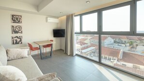 A modern living room with a large window, a flat-screen TV, a sofa, and a small table with chairs. - Sky-High Central Penthouse w. Stunning Views - 01

 (Chania, Crete)