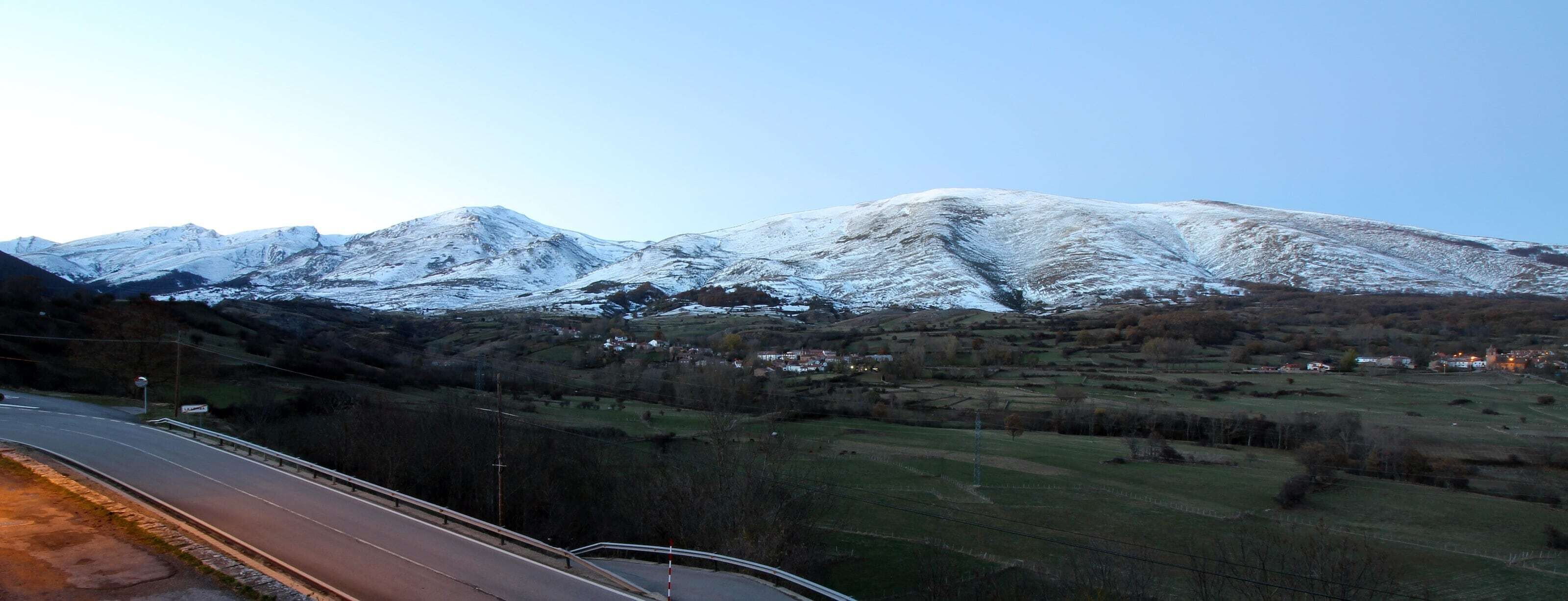 Un paisaje montañoso con picos nevados, una carretera y edificios dispersos en el valle.