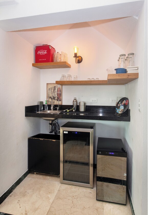Een compacte keuken met een spoelbak, een vaatwasser en een compacte koelkast. Boven het aanrecht staan planken met glazen en een rode Coca-Cola doos.