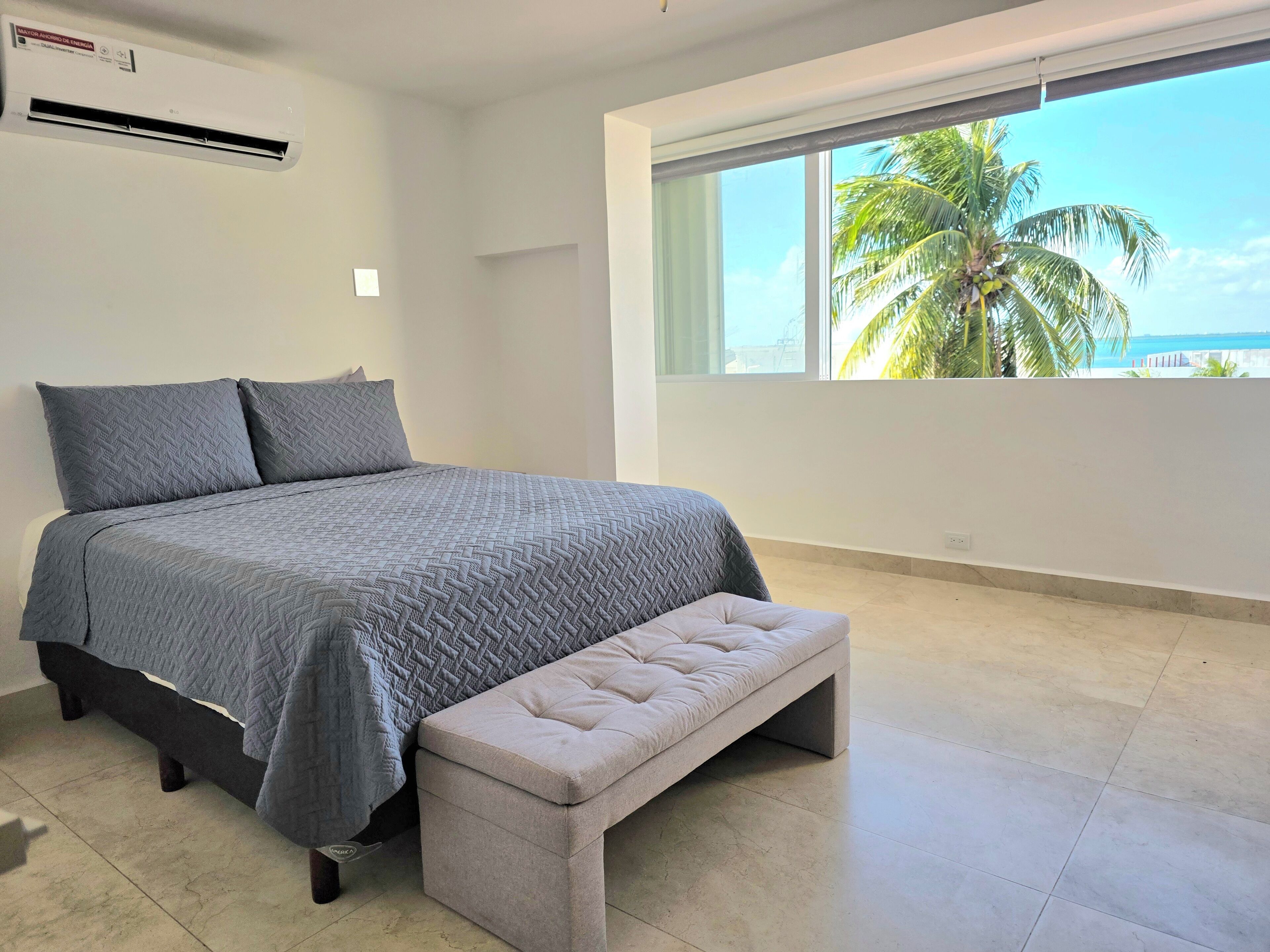 A bedroom with a bed, a bench, a window with a view of palm trees, and an air conditioning unit.