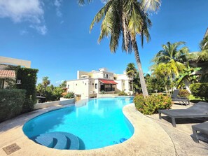 A swimming pool with lounge chairs, surrounded by palm trees and a building with a red roof.