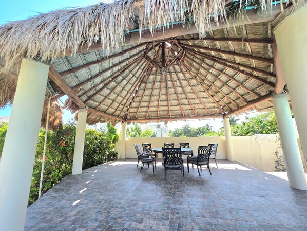 A covered outdoor seating area with stone flooring and thatched roof, surrounded by greenery.