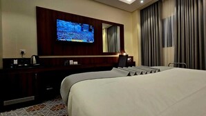 Ein Hotelzimmer mit einem großen Bett, einem an der Wand befestigten Fernseher, einem Schreibtisch mit Computer und einem Fenster mit Vorhängen.