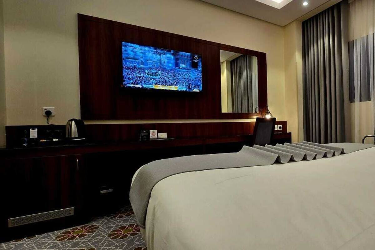 A hotel room with a large bed, a TV mounted on the wall, a desk with a computer, and a window with curtains.