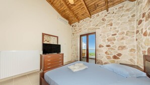 Une chambre avec un mur en pierre, un plafond en bois, un lit avec une literie bleue, un téléviseur posé sur une commode en bois et un balcon avec vue.