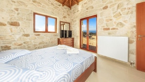 Une chambre avec des murs en pierre, un plafond en bois, un lit recouvert d’une couvre-lit à motifs, une commode, un téléviseur et une vue sur la mer par une porte vitrée.