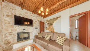 Un salon avec un mur en pierre, un plafond en bois, un téléviseur à écran plat, une cheminée, un canapé avec des coussins à rayures et un escalier menant à une autre pièce.