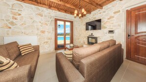 Un salon chaleureux avec des murs en pierre, un plafond en bois, une cheminée et une vue sur la mer par une porte vitrée.