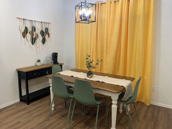 A dining area with a wooden table, green chairs, a white table runner, a black vase with a plant, a coffee maker, and a wall hanging.