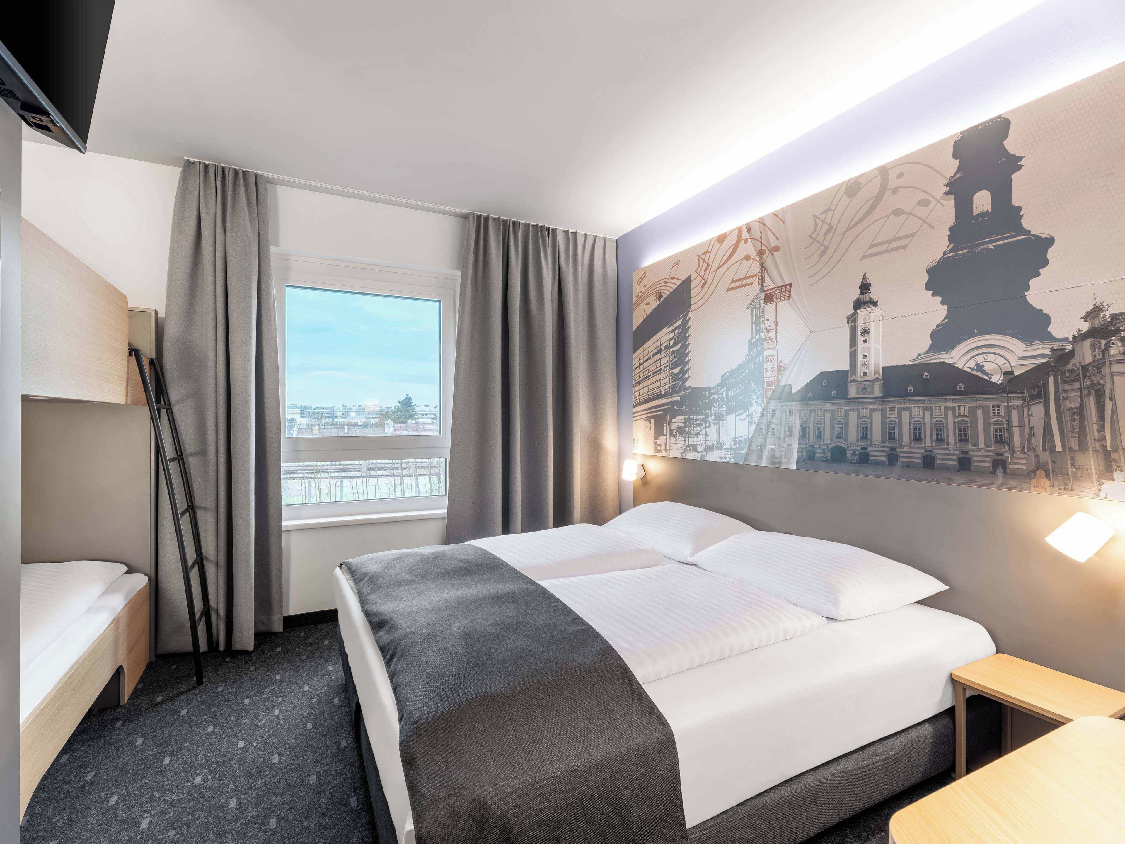 A hotel room with a bed, a desk, a chair, a window with curtains, and a wall with a cityscape mural.