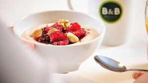 A bowl of yogurt with berries and nuts, a spoon, and a mug with a logo. - B&B HOTEL St. Pölten (St. Pölten)