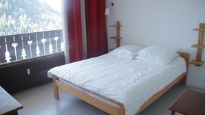 Un dormitorio con una cama de madera, un ventanal con vistas a un bosque, una cortina roja y una mesita.