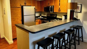 A kitchen with wooden cabinets, a stainless steel refrigerator, a microwave, and a stove. There are four black bar stools at the island. - SkiBoltonValleyVTPresWkFeb12-19Slopeside1bdrm2bthSteSlps8 (Richmond)