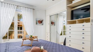 Un dormitorio con cama, televisor, mesita de noche de madera y vista a un edificio.