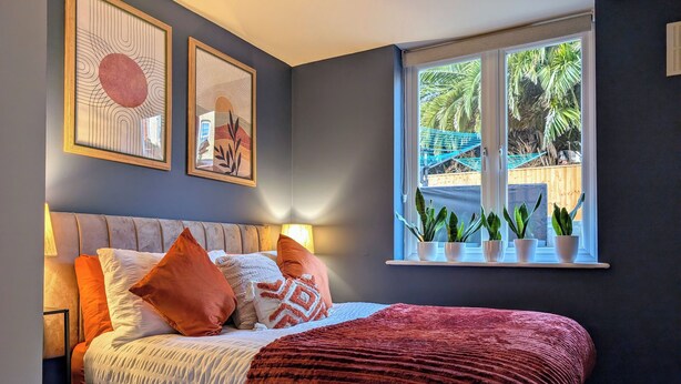 Ein Schlafzimmer mit einem Bett, Kissen, einer roten Decke und einem Fenster mit Pflanzen.