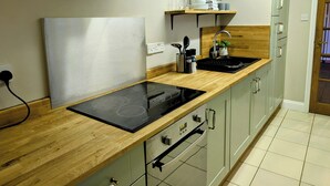 Eine moderne Küche mit einer schwarzen Induktionskochplatte, einem eingebauten Backofen und einem Edelstahlspülbecken. Darüber befinden sich Holzregale mit Geschirr und Gläsern.